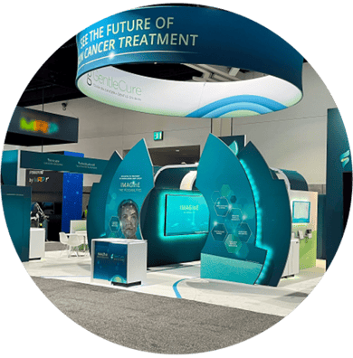 Trade show booth showcasing the future of cancer treatment. The booth features a teal color scheme, a large banner with the text "See the Future of Cancer Treatment," and a prominent display with the logo "Gentle Cure. Trade show booth showcasing the future of cancer treatment. The booth features a teal color scheme, a large banner with the text "See the Future of Cancer Treatment," and a prominent display with the logo "Gentle Cure.